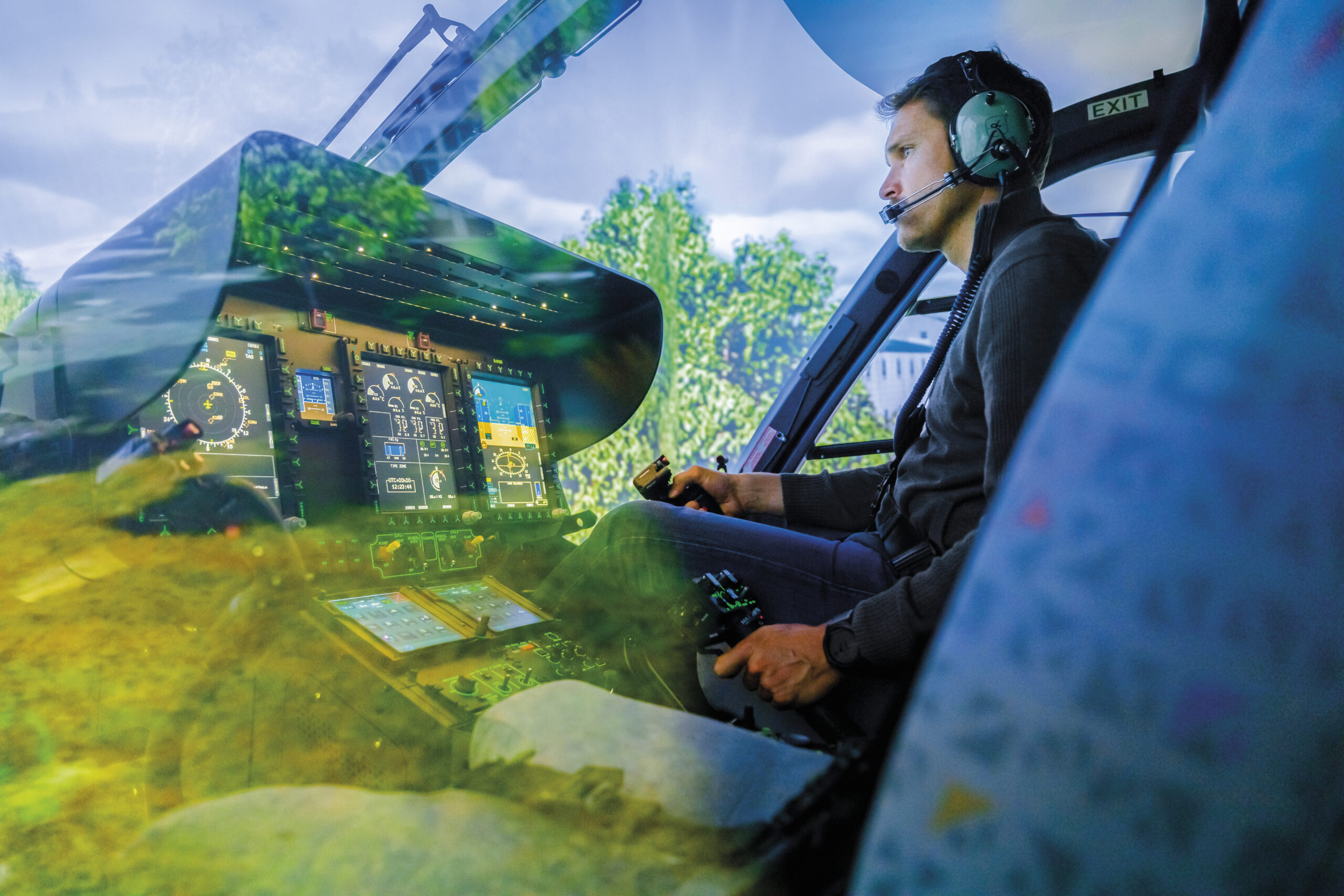 A man wearing a headset with a microphone is seated in a helicopter simulator, looking focused as he operates the controls. The immersive display screens show a realistic green landscape with trees and a cloudy sky. The cockpit's instrument panel is illuminated with various flight data