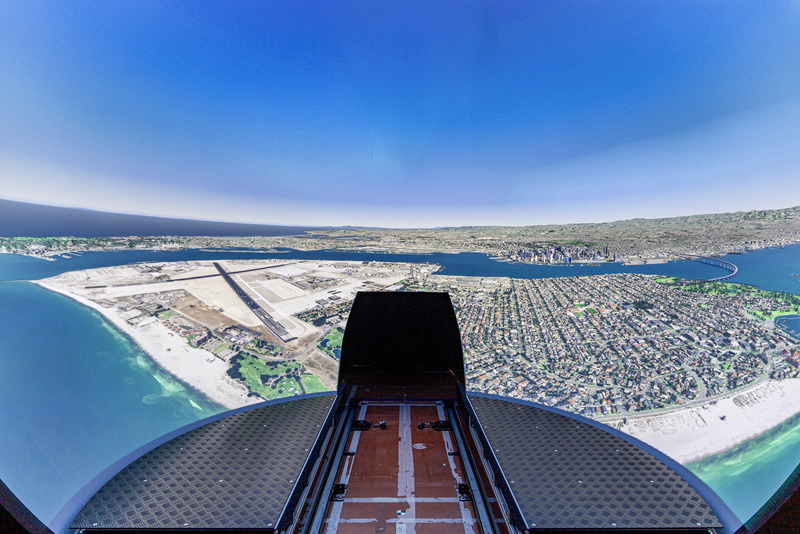 View from the cockpit position into the 220° projection dome, showing simulated aerial imagery of a coastal city and runway